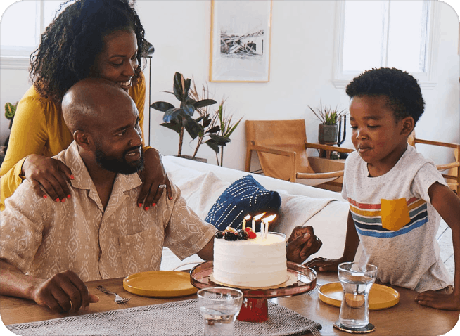 A smiling husband and wife sitting at their living room table with their son as he blows out the candles on his birthday cake.